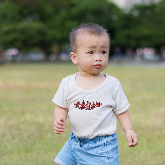 Child wearing a white t-shirt with Nola Til Ya Die Crawfish Krewe and blue shorts standing in a grassy field.
