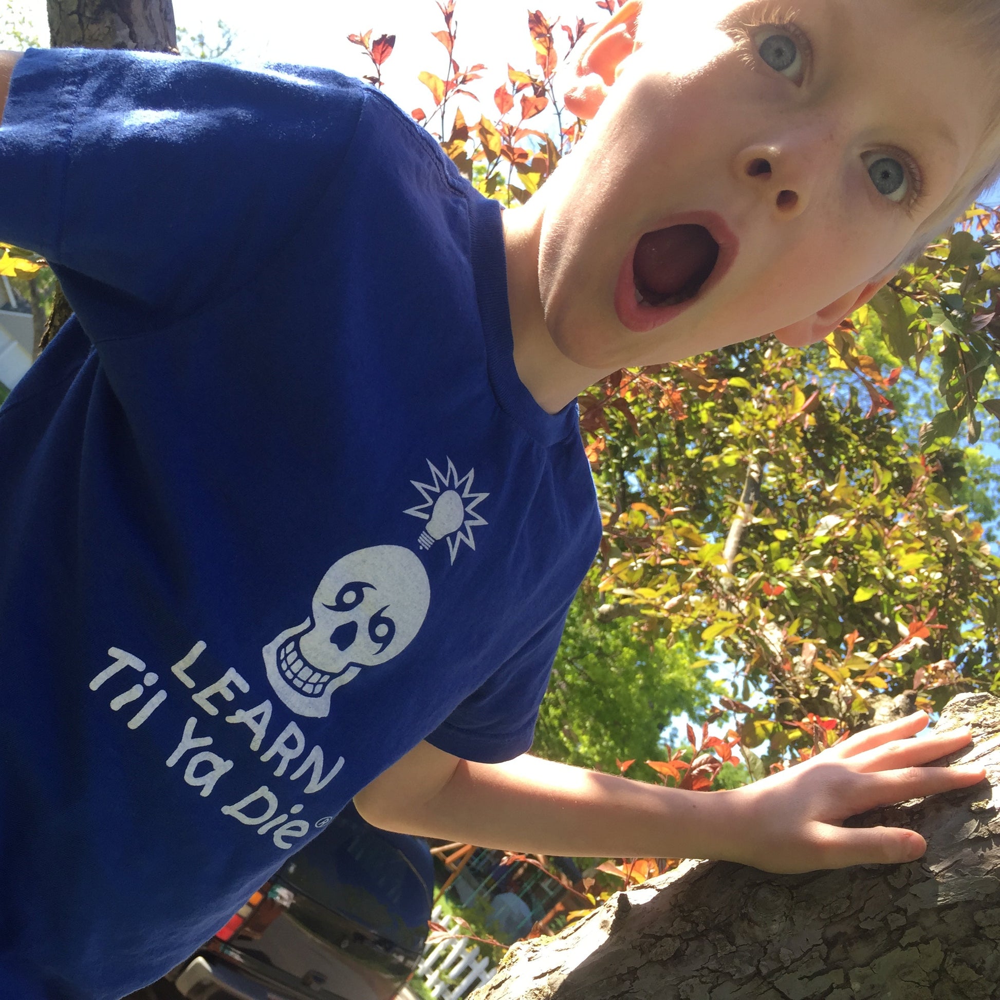 A young boy wearing a blue t-shirt with a white Nola Til Ya Die skull and the text 'LEARN TIL YA DIE' looking up with a surprised expression outdoors.