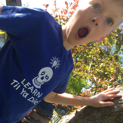 A young boy wearing a blue t-shirt with a white Nola Til Ya Die skull and the text 'LEARN TIL YA DIE' looking up with a surprised expression outdoors.