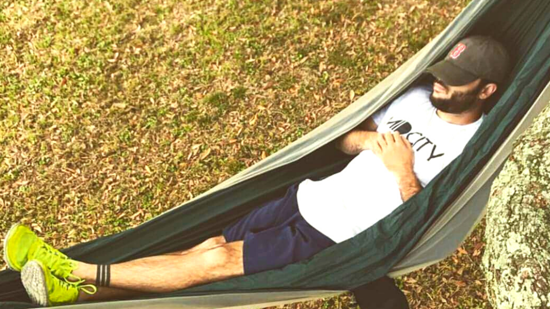 A man relaxes in a green hammock strung between trees outdoors on grassy ground. He is wearing a Nola Til Ya Die white t-shirt with Mid City Til Ya Die, navy shorts, neon green sneakers, and a dark cap.