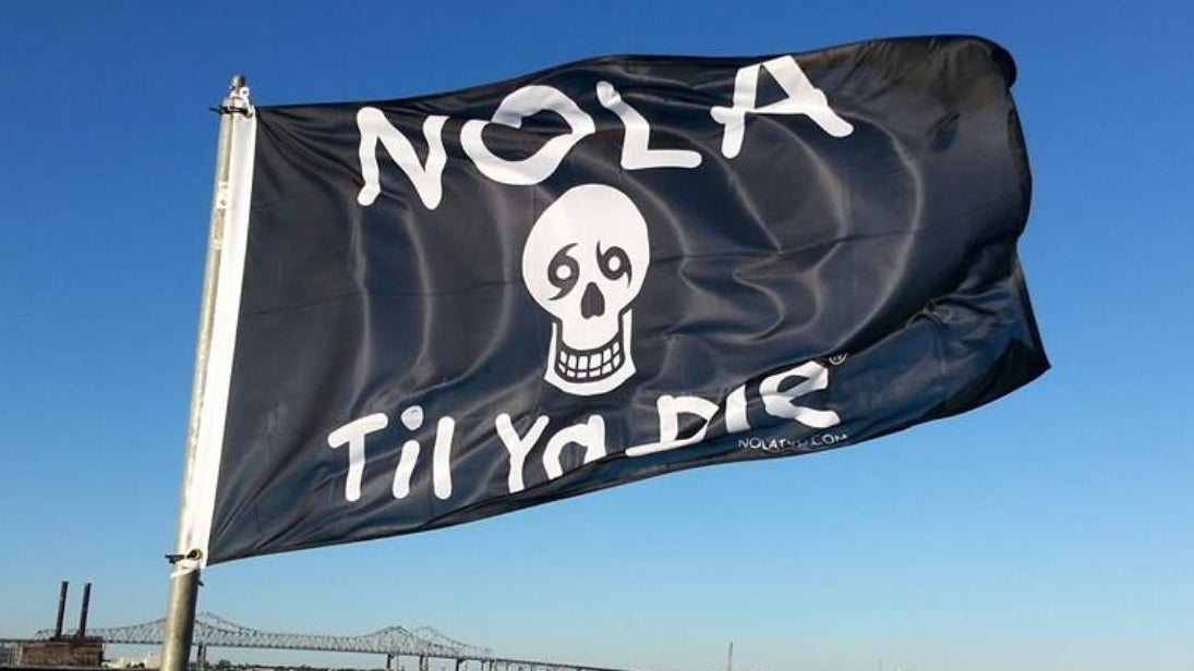 Nola Til Ya Die flag with white text and skull design flying over the Mississippi River in a clear blue sky.