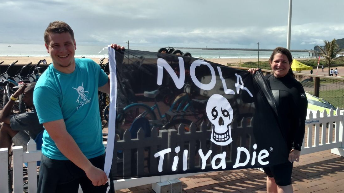 Two people standing on a beachside deck in South Africa holding a black flag that reads 'NOLA Til Ya Die' with a white skull graphic and a man guy wearing a Surf Til Ya Die t-shirt in aqua blue.