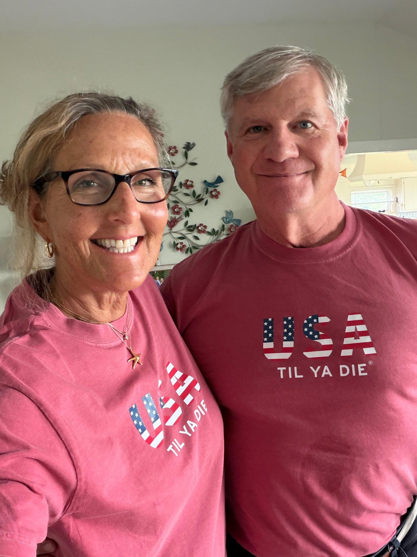A smiling couple indoors, both wearing matching  t-shirts with a distressed 'USA TIL YA DIE' graphic featuring an American flag pattern from Nola Til Ya Die.